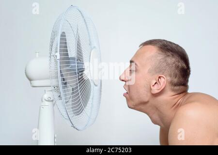 L'uomo con la barba stoppia soffre di calore e cerca di raffreddarsi vicino al ventilatore. Foto Stock