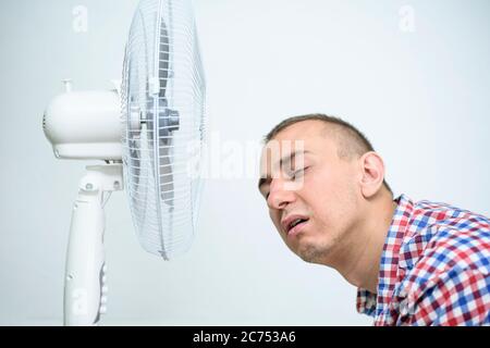L'uomo con la barba stoppia soffre di calore e cerca di raffreddarsi vicino al ventilatore. Foto Stock