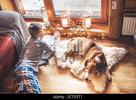 Libro di lettura del ragazzo sul pavimento vicino a scivolare il suo cane di aquila su pelle di pecora in atmosfera accogliente casa. Momenti tranquilli di accogliente casa concetto immagine. Foto Stock