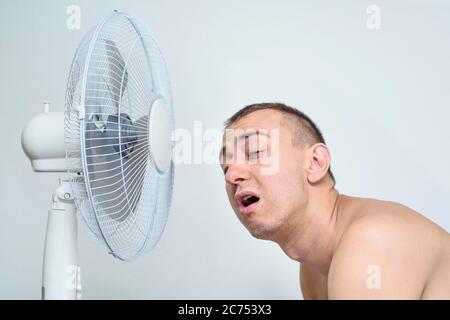 L'uomo con la barba stoppia soffre di calore e cerca di raffreddarsi vicino al ventilatore. Foto Stock