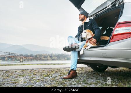 Uomo impanato in abiti caldi seduto con beagle nel bagagliaio dell'auto. Viaggiare con l'immagine concettuale degli animali domestici Foto Stock
