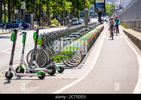 I ciclisti si trovano in bicicletta su una pista ciclabile, accanto a una flotta di biciclette condivise Velib, allineate a una stazione di attracco insieme agli scooter elettrici Lime a Parigi. Foto Stock