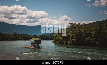 Casa di legno del fiume Drina che si erge su una roccia nel mezzo del fiume. Dietro ci sono montagne e cielo blu con nuvole in una giornata di sole, immagine orizzontale Foto Stock