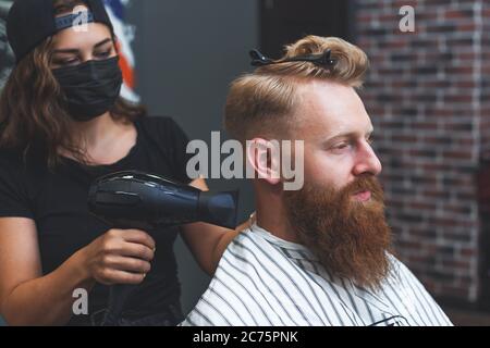 Il barbiere femminile in maschera asciuga i capelli al cliente con un asciugacapelli durante il taglio dei capelli. La distanza sociale nei luoghi pubblici. Foto Stock