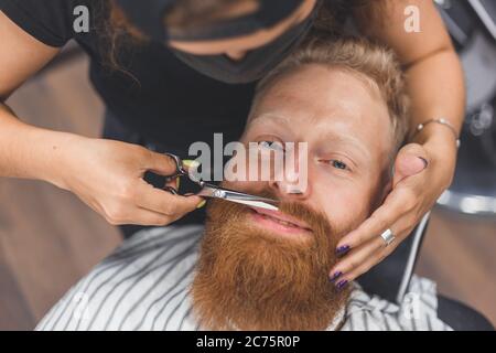 Un uomo in un barbiere. Baffi da taglio per barbiere donna. Barbiere donna in maschera. Foto Stock