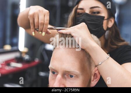 Il barbiere delle donne fa il taglio di capelli piatto con le forbici un uomo bearded. Servizio in una maschera da virus. Foto Stock