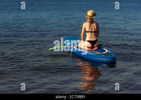 Giovane donna attraente seduta su tavola da paddle, SUP. Concetto di vita attiva, sport, attività per il tempo libero. Donna caucasica a bordo di viaggio in estate sera. Vacanza, resort, divertimento. Ritagliato. Foto Stock