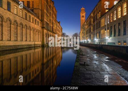 Salts Mill, il Sito Patrimonio Mondiale dell'UNESCO in Saltaire, si illumina in arancione durante le ore di colore blu con l'architettura storica si riflette nel canale. Foto Stock