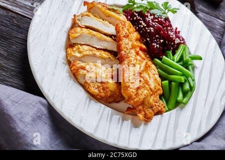 Schnitzel di pollo servito con insalata di barbabietole e fagiolo verde croccante al vapore su un piatto bianco su un tavolo di legno scuro, vista orizzontale dall'alto, vicino- Foto Stock