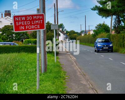 Un cartello rosso avverte gli automobilisti di un nuovo limite di velocità di 30 mph in vigore nel villaggio rurale di Halsall nel Lancashire, Regno Unito. Un'auto blu passa sulla destra. Foto Stock