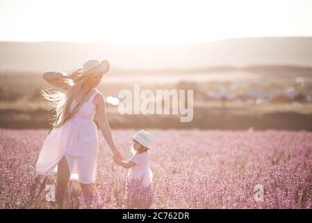 Giovane donna sorridente che tiene la bambina di 1-2 anni che cammina in prato di lavanda all'aperto sopra la luce del sole sullo sfondo. Stagione estiva. Messa a fuoco selettiva. Foto Stock