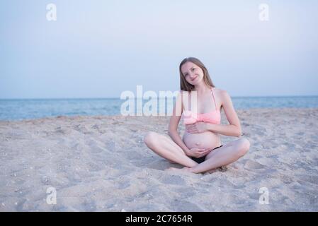 Donna incinta siede sulla spiaggia e gode. Luce soffusa da sera, spiaggia deserta. Foto Stock
