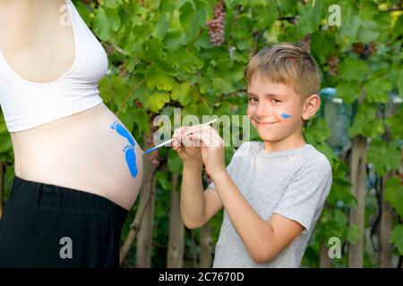 Il bambino felice disegna con una spazzola sul ventre dell'impronta della sua mamma incinta. Concetto di gravidanza. Primo piano Foto Stock