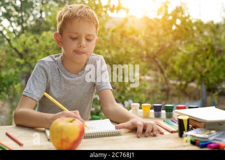 Il ragazzo carino disegna con matite ancora vita. Aria aperta. Giardino sullo sfondo. Concetto creativo. Foto Stock