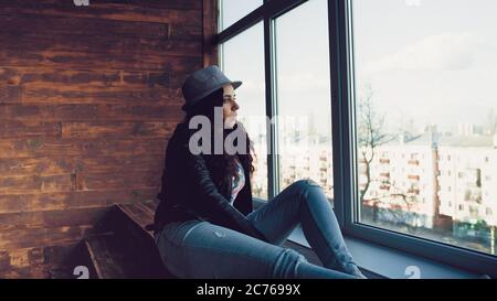 Giovane bella donna in cappello e giacca di pelle seduta da finestre. Ritratto di bella donna che guarda fuori la finestra di giorno. Foto Stock