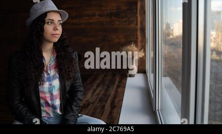 Giovane bella donna in cappello e giacca di pelle seduta da finestre. Ritratto di bella donna che guarda fuori la finestra di giorno. Foto Stock