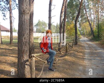 Primo piano ritratto di una donna anziana sorridente che si trova su una recinzione di legno vicino alla strada provinciale nella pineta. Foto Stock