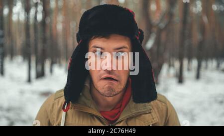 Uomo grimaces in foresta nella stagione invernale. Ritratto di pazzo maschio che indossa copricapo e grimacing faccia da vicino nella foresta invernale. Foto Stock