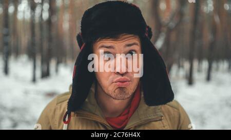 Uomo grimaces in foresta nella stagione invernale. Ritratto di pazzo maschio che indossa copricapo e grimacing faccia da vicino nella foresta invernale. Foto Stock