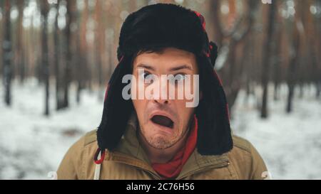 Uomo grimaces in foresta nella stagione invernale. Ritratto di pazzo maschio che indossa copricapo e grimacing faccia da vicino nella foresta invernale. Foto Stock