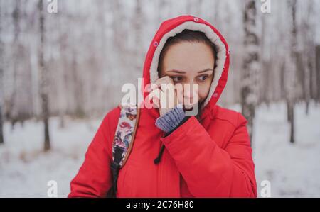 Giovane donna in giacca rossa e cappuccio sfrega l'occhio nella foresta invernale. Primo piano. Foto Stock