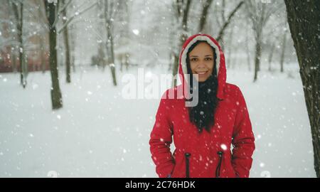 Ritratto di giovane donna in giacca rossa e cappuccio nella stagione invernale. Bella donna sorride, gioendo in caduta di neve. La neve soffice avvolge tutto Foto Stock