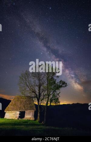 Vecchia casa colonica abbandonata fienile con due alberi accanto ad esso sparato di notte contro un cielo stellato con un nucleo galattico modo latteo visto sopra sparato a Dumesti, SA Foto Stock