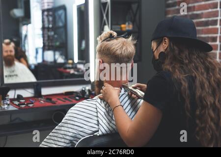 Il barbiere femminile in maschera taglia i capelli di un uomo con il regolacapelli. Acconciatura durante la distanza sociale Foto Stock