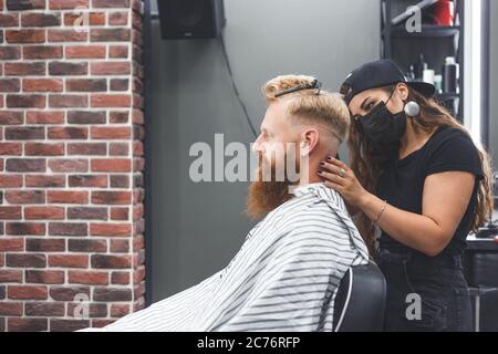 Il barbiere femminile in maschera taglia i capelli di un uomo con il regolacapelli. Acconciatura durante la distanza sociale Foto Stock