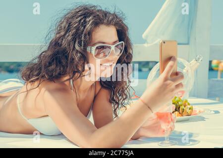 Closeup giovane bella felice, eccitato donna sorridente guardando sul cellulare prendere selfie, ascoltare musica all'aperto sul lettino al mare lago ba Foto Stock