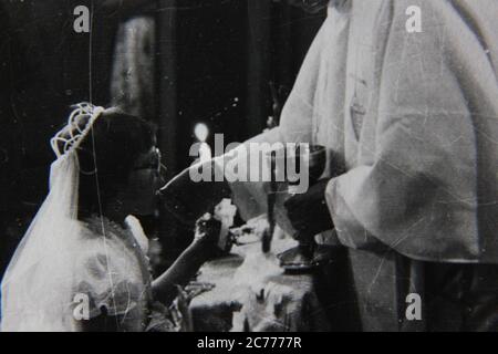 Fine anni '70 vintage nero e bianco stile di vita fotografia di una giovane ragazza che riceve la sua prima comunione Santa da un sacerdote in chiesa. Foto Stock