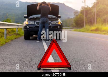 Cartello rosso triangolo su strada per l'avvertenza: Avere un'auto con cofano aperto e uomo che ripara un'auto. Foto Stock
