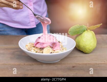 Le donne versano la fragola dello yogurt dal cucchiaio al recipiente e nella ciotola hanno il vetrino alla banana, l'avena guava sul tavolo di legno Foto Stock