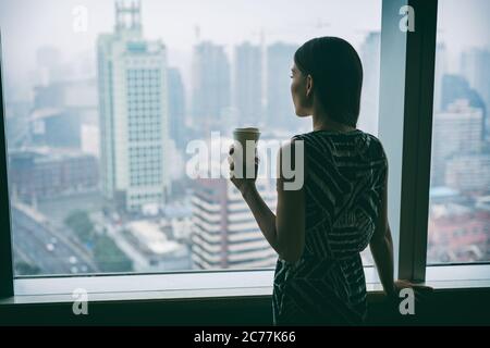 Donna d'affari che beve caffè al lavoro contemplativo guardando fuori dalla finestra dell'alto grattacielo edificio durante la pausa del tè del mattino. Stress, mentale Foto Stock