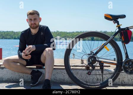 Ritratto di un atleta di ciclista che riposa dopo un giro in bicicletta usando uno smartphone in mano. All'aperto. Sport primaverili. Il ragazzo ha venticinque anni Foto Stock
