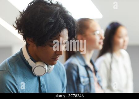 Primo piano di un serio studente di razza mista con cuffie wireless intorno al collo seduto in fila di compagni di gruppo alla conferenza universitaria Foto Stock