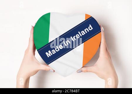 Iscrizione fatta in Irlanda, la bandiera dell'Irlanda. Mani femminili che tengono una scatola a forma di cuore. Sfondo bianco. Foto Stock