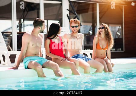 Quattro giovani adulti seduti vicino alla piscina con le gambe in acqua Foto Stock