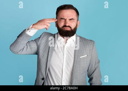 Uomo d'affari sottolineato che punta le dita alla testa come una pistola, che vuole sparare su sfondo blu Foto Stock