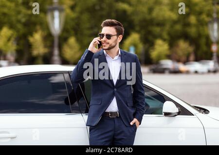 Maganer finanziario serio in abbigliamento formale che parla sul cellulare vicino alla sua auto a grande città Foto Stock