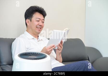 Un uomo asiatico di mezza età che legge nel soggiorno con un purificatore d'aria acceso. Foto Stock