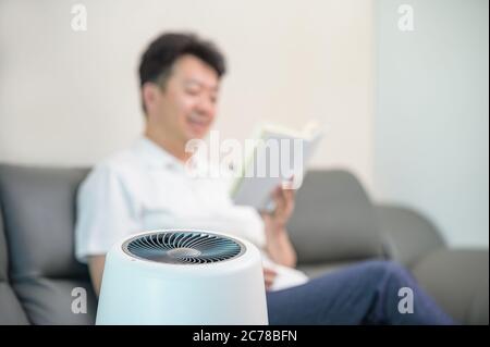 Un uomo asiatico di mezza età che legge nel soggiorno con un purificatore d'aria acceso. Sfocatura dello sfondo. Foto Stock