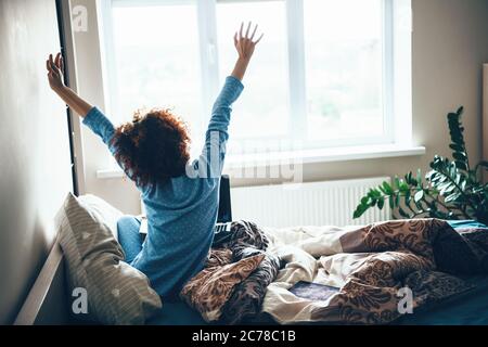 Donna caucasica riccia capelli in pajama blu che si allunga a letto la mattina presto Foto Stock
