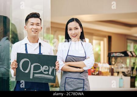 Sorridente giovane proprietario di cafe' Vietnamita con un cartello aperto in piedi all'entrata del cafe e che accoglie i clienti all'interno Foto Stock