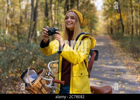 Fotografo che tiene una fotocamera retrò, scattando foto nel parco. Bella donna sorridente con impermeabile giallo che cammina nella foresta autunnale. Viaggi Foto Stock