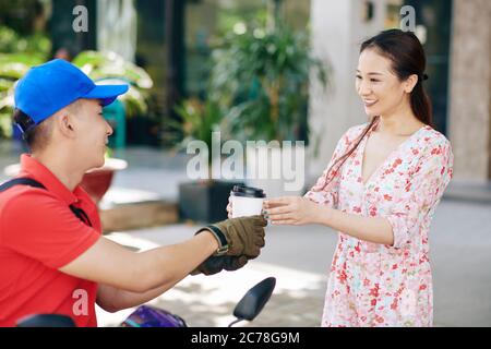 Uomo di consegna che dà una tazza di caffè da portare via a sorridente bella donna vietnamita Foto Stock