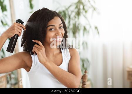 Acconciatura. Sorridente Donna nera applicando lo spray testurizzante ai suoi bei capelli ricci Foto Stock