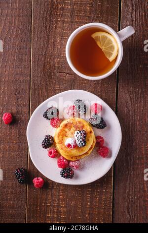 Frittelle di formaggio cottage, frittelle di cagliata dessert con lampone e frutti di bosco in piatto vicino alla tazza di tè caldo con fetta di limone Foto Stock