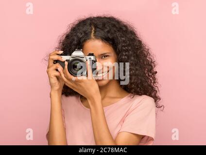 Felice attraente giovane donna nera con una fotocamera vintage che si concentra sullo spettatore con un sorriso amichevole e luminoso sullo sfondo di uno studio rosa Foto Stock