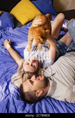 Un autentico colpo di giovane coppia allegra che si trova nel letto e gioca con il loro gatto zenzero. Luce naturale del sole, concetto di fine settimana a casa Foto Stock
